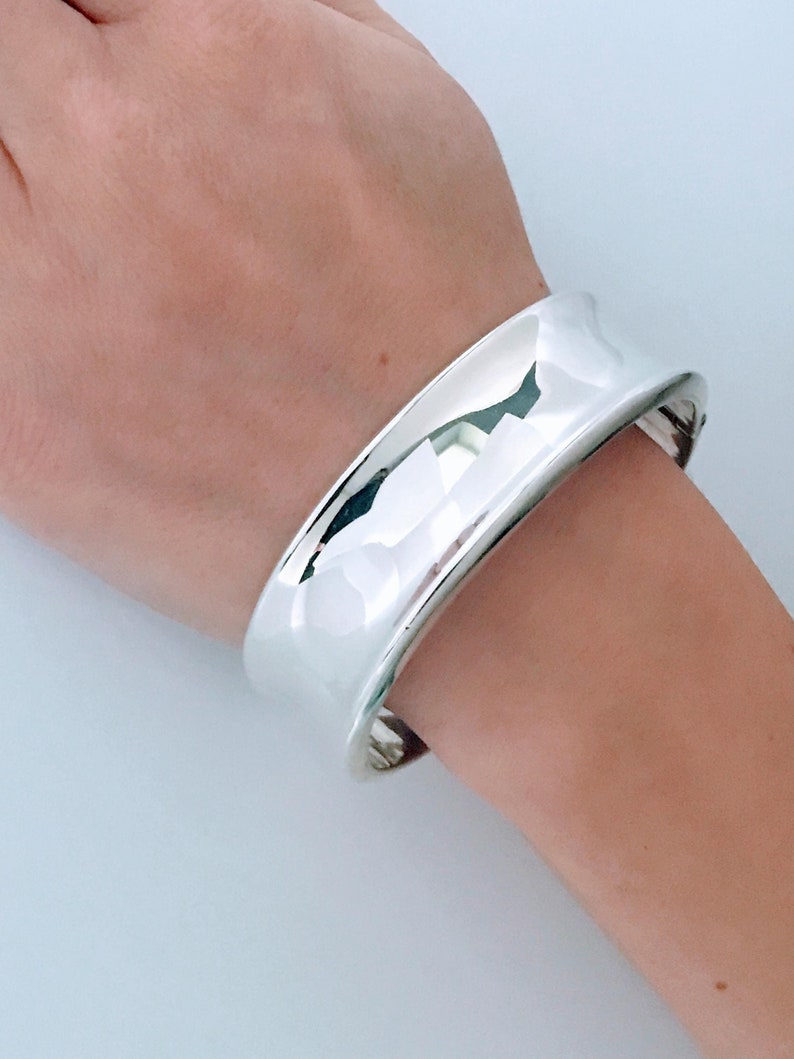 May include: A silver bangle bracelet with a smooth, rounded surface. The bracelet is wide and has a simple, modern design.