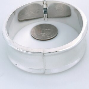 May include: A silver hinged bangle bracelet with a smooth, rounded surface. The bracelet is open and a US quarter is placed inside for size reference.