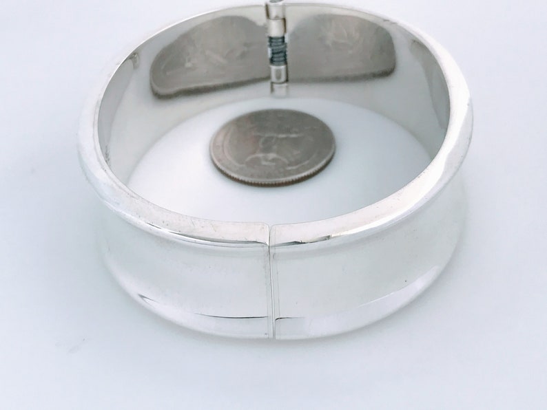 May include: A silver hinged bangle bracelet with a smooth, rounded surface. The bracelet is open and a US quarter is placed inside for size reference.