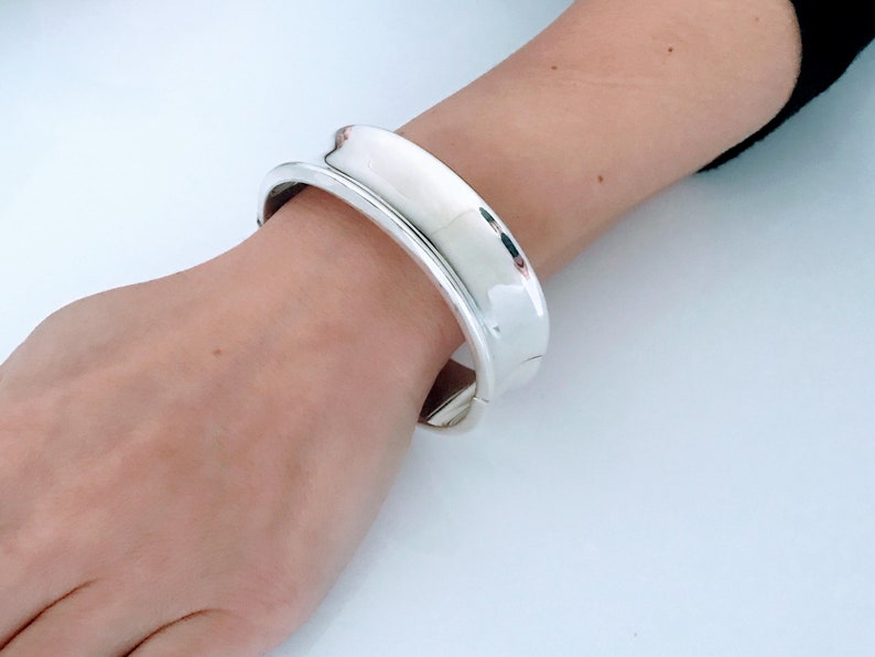 May include: A silver bangle bracelet with a wide, smooth, rounded design. The bracelet is open and has a hinged closure.