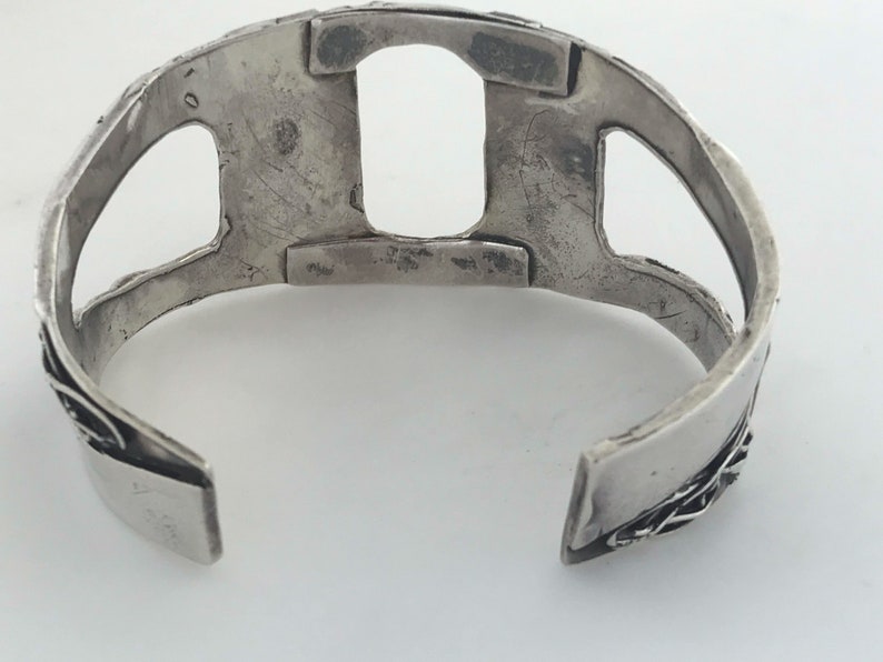 May include: Silver bracelet with a cutout design. The bracelet is open on one side and has a textured surface.