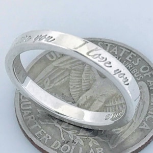 May include: A silver band ring with the inscription "I love you" on the outside of the band. The ring is resting on top of a US quarter.