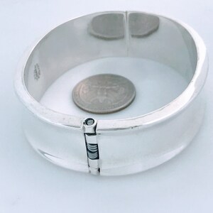 May include: A silver hinged bangle bracelet with a polished finish. The bracelet is open and a US quarter is placed inside to show the size.