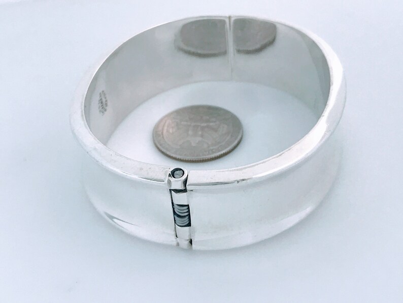 May include: A silver hinged bangle bracelet with a polished finish. The bracelet is open and a US quarter is placed inside to show the size.
