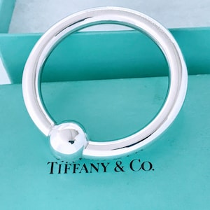 May include: A polished silver bracelet with a spherical accent, resting on a Tiffany & Co. box. The bracelet is a circular design with a smooth, reflective surface. The box is a light blue color, with the brand name printed in black.