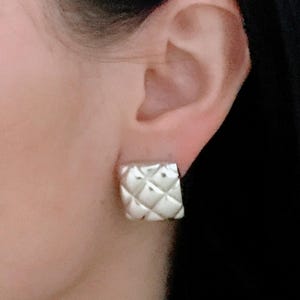 May include: A silver square earring with a quilted design is shown. The earring is a stud style and is worn on the ear. The earring has a shiny finish and a geometric pattern.
