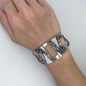 May include: A silver cuff bracelet with a geometric design. The bracelet has a series of open rectangles with intricate details.