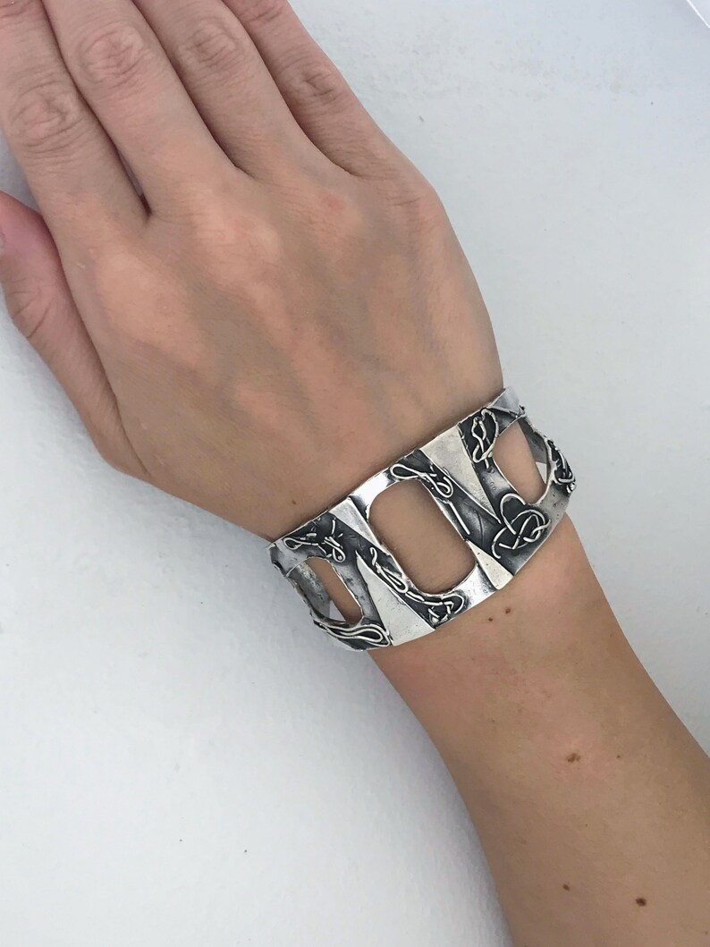 May include: A silver cuff bracelet with a geometric design. The bracelet has a series of open rectangles with intricate details.