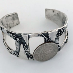 May include: A silver bracelet with a textured, geometric design. The bracelet is open and has a gap in the middle. A US quarter is placed in the gap to show the size of the bracelet.