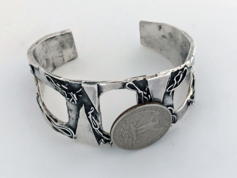 May include: A silver bracelet with a textured, geometric design. The bracelet is open and has a gap in the middle. A US quarter is placed in the gap to show the size of the bracelet.
