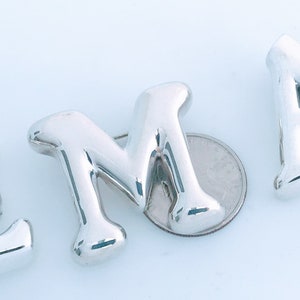 May include: Silver letter charms in the shape of the letters E, M, and A. The charms are shiny and have a smooth, rounded surface.