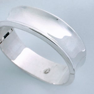 May include: A silver bangle bracelet with a smooth, curved design. The bracelet is open and has a hinged clasp.