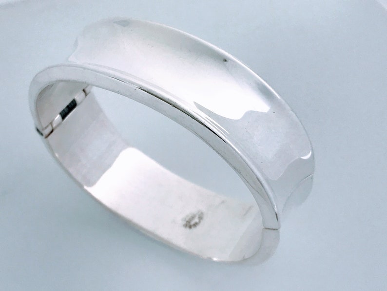 May include: A silver bangle bracelet with a smooth, curved design. The bracelet is open and has a hinged clasp.