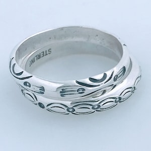 May include: Two silver rings with intricate engraved designs. The top ring has a wider band and features a more elaborate pattern. The bottom ring has a narrower band and a simpler pattern. The inscription "STERLING" is visible on the inside of the top ring.