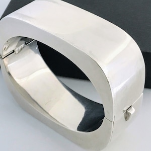 May include: A polished silver bracelet with a rectangular, curved design. The hinged cuff bracelet has a secure clasp and a smooth, reflective surface. The jewelry piece is displayed on a white surface with a black box in the background.
