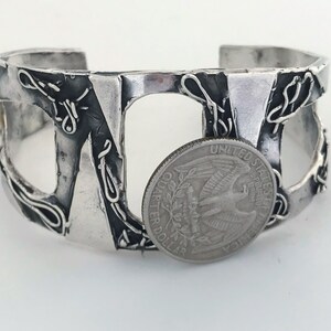May include: A silver cuff bracelet with an abstract design. The bracelet is made of thick, textured metal and has a cutout design. A US quarter is placed next to the bracelet for size reference.