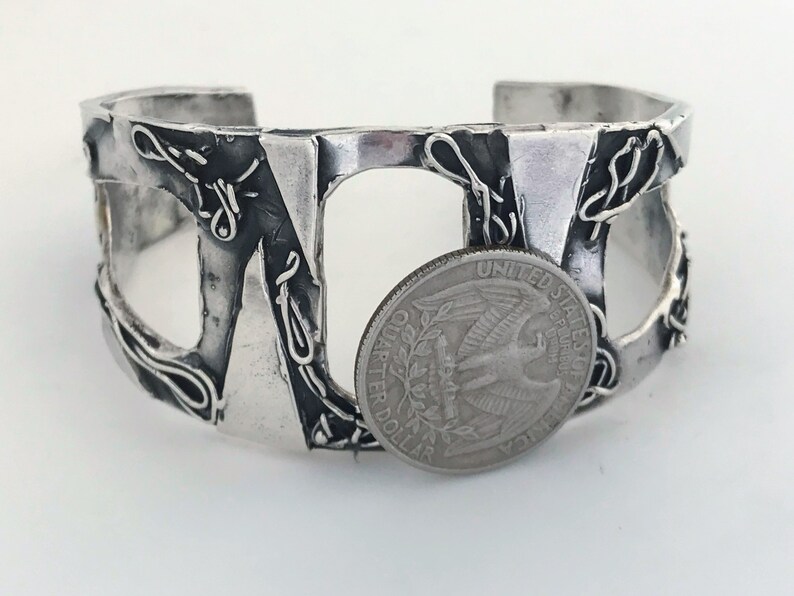 May include: A silver cuff bracelet with an abstract design. The bracelet is made of thick, textured metal and has a cutout design. A US quarter is placed next to the bracelet for size reference.