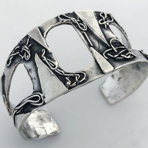 May include: A silver cuff bracelet with a geometric design and intricate details. The bracelet is made of metal and has a textured surface.