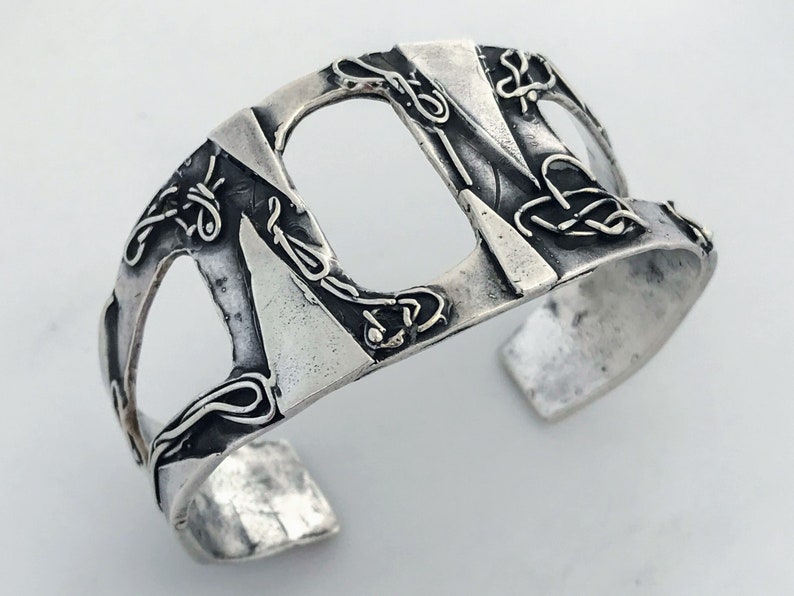 May include: A silver cuff bracelet with a geometric design and intricate details. The bracelet is made of metal and has a textured surface.