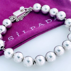 May include: A silver bracelet featuring large, polished spherical beads. The bracelet has a rectangular clasp and a small chain. The bracelet is displayed on a purple cloth bag with the brand name "SILPADA" printed on it.