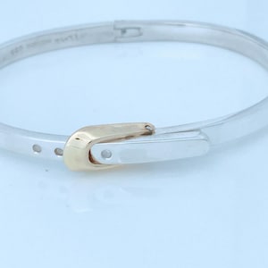 May include: A silver-toned hinged bangle bracelet featuring a polished finish and a gold-toned buckle-style clasp mechanism. The bracelet has a smooth, rounded band design with a secure closure, showcasing a two-tone metallic aesthetic.