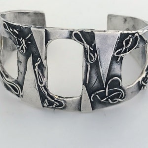 May include: A silver cuff bracelet with a geometric design. The bracelet is made of textured metal and has a unique, abstract pattern.