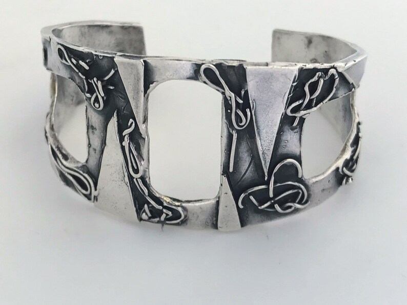 May include: A silver cuff bracelet with a geometric design. The bracelet is made of textured metal and has a unique, abstract pattern.