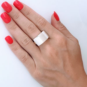May include: A silver ring with a wide, flat band. The ring is worn on the middle finger of a hand with red nail polish.