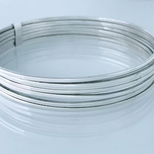 May include: A stack of silver-colored bangles. The bracelets are thin and flat, with a smooth, reflective surface. The bangles are stacked together, creating a layered effect. The overall aesthetic is minimalist and modern.
