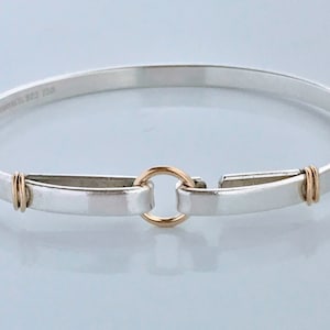 May include: A silver bangle bracelet with a gold clasp and accents. The bracelet has a simple, elegant design with a smooth, polished finish. The clasp is a circular gold ring that connects the two ends of the bracelet. The bracelet is approximately 7 inches in circumference.