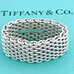May include: A silver ring featuring a woven chain-link design. The interior of the band is engraved with the text Tiffany & Co. 925. The ring is positioned against a bright turquoise background with the text Tiffany & Co. printed in dark blue.