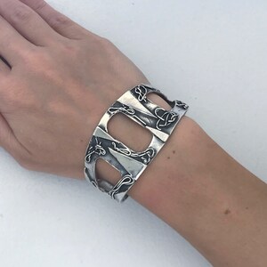 May include: A silver cuff bracelet with an intricate design of interwoven lines and shapes. The bracelet is open on one side and has a wide, flat band.