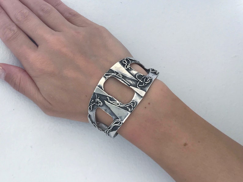 May include: A silver cuff bracelet with an intricate design of interwoven lines and shapes. The bracelet is open on one side and has a wide, flat band.