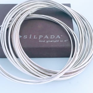 May include: A set of silver bangle bracelets stacked on top of each other. The bracelets are all the same width and have a smooth, polished finish. The bracelets are in a box with the text "SílPada. find yourself in it" printed on the box.