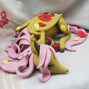 Puede incluir: Una escultura de arcilla polimérica hecha a mano de un dragón durmiendo con pelo rosa, escamas verdes y marrones y decoraciones de otoño como hojas y flores.