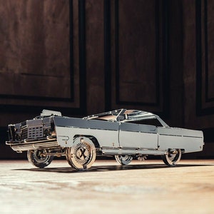 May include: A silver model car with a detailed design. The car has a classic body style with a long bonnet and a chrome bumper. The car is sitting on a wooden surface.