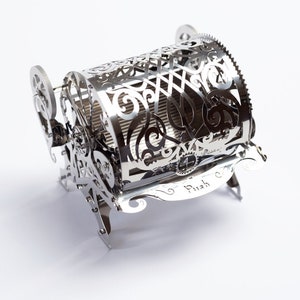 May include: A silver metal sculpture of a small, intricate, ornate, mechanical contraption with a wheel and gears. The sculpture is on a white background. The sculpture has the words "To Push" engraved on it.