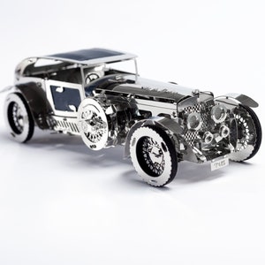 May include: A silver metal model car with intricate details, including a detailed engine and wheels. The car has a black roof and a number plate that reads "TUD5".