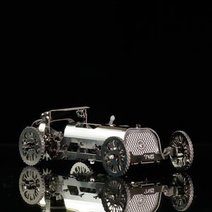 May include: A silver metal model of a classic racing car, with intricate detailing on the wheels and body. The car is displayed on a reflective surface against a black backdrop, showcasing its design.