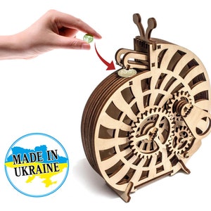 May include: Wooden snail shaped coin bank with a gear mechanism. The bank is made of light brown wood and has a slot on the top for inserting coins. The coin bank is made in Ukraine, as indicated by the label on the bottom.