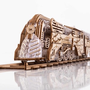 May include: A wooden model of a vintage steam locomotive train with intricate details. The model is made of light brown wood and features a detailed engine, wheels, and a passenger car. The train is on a track and is ready to be assembled.