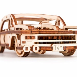 May include: A detailed wooden model car, featuring visible gears and a license plate with the text "T4M". The car is made from light-coloured wood with darker accents, and has a classic car design. The model is approximately 20 cm in length.