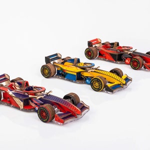 May include: Three wooden Formula 1 race cars in red, blue, and yellow. The cars have detailed designs, including visible engine components and sponsor logos on the tyres. The cars are set against a white background.