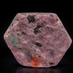 May include: A hexagonal-shaped, rough-cut ruby gemstone. The stone is a deep pink color with dark green and black inclusions. The surface has a slightly textured appearance, and the gemstone is displayed against a black background.
