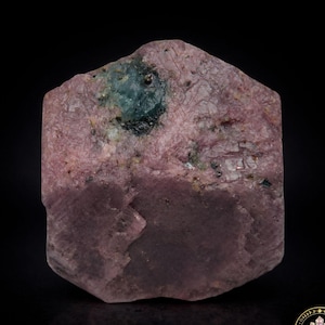 May include: A rough-cut, natural ruby gemstone with a deep pink hue. The stone has a textured surface with areas of darker pink and a patch of teal-colored inclusions. The gemstone is displayed against a black background.