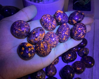 May include: A collection of polished sodalite stones, each with a dark purple base and vibrant orange speckles. The stones are egg-shaped and held in a hand, with more stones scattered below. The lighting gives the stones a glowing appearance.