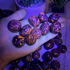 May include: A collection of polished sodalite stones, each with a dark purple base and vibrant orange speckles. The stones are egg-shaped and held in a hand, with more stones scattered below. The lighting gives the stones a glowing appearance.