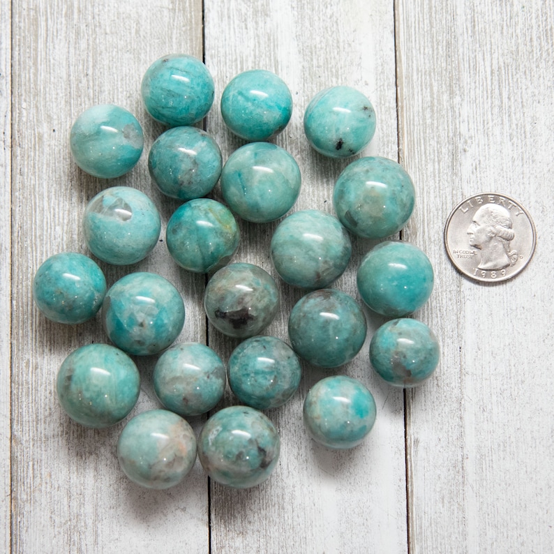 May include: A collection of polished, turquoise-colored spheres, possibly gemstones, are scattered on a white wooden surface. A U.S. quarter coin is included for size reference. The spheres vary slightly in color and pattern, with some showing darker inclusions.