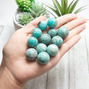 May include: A hand holds a collection of polished, spherical Amazonite gemstones. The stones are a mix of teal and light blue with subtle gray and brown inclusions. The gemstones are smooth and reflect light, suggesting a polished finish.