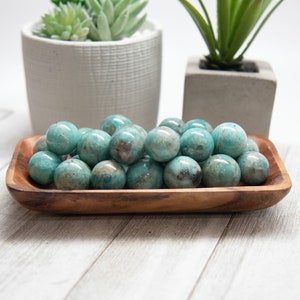 May include: A wooden bowl filled with polished, teal-colored spheres. The spheres have a marbled appearance with darker spots. Two small potted plants are in the background. The bowl is made of a light brown wood.